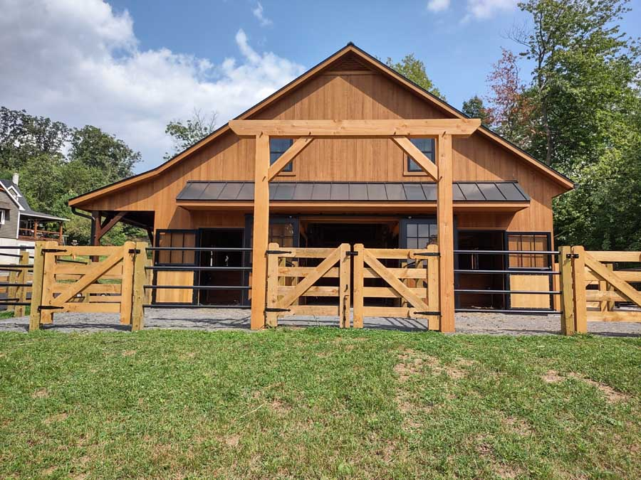 A wooden horse barn with a matching timber frame gate and fence, set against a bright, sunny landscape.