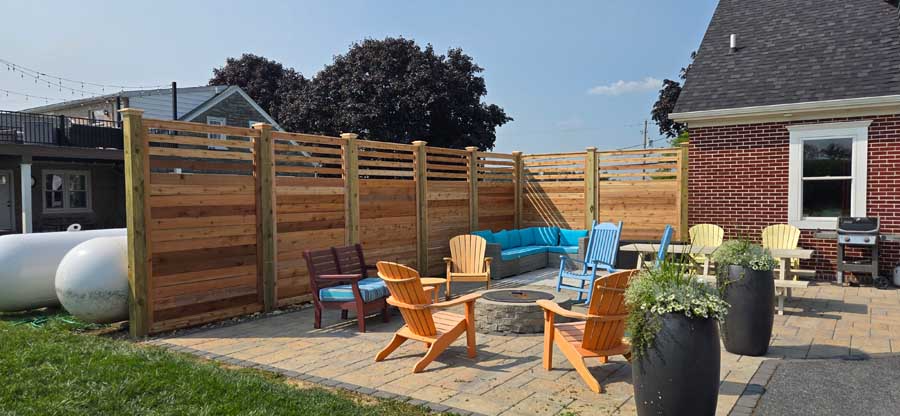 Patio area with wood privacy fence, fire pit, Adirondack chairs, and outdoor seating on a stone-paved ground.