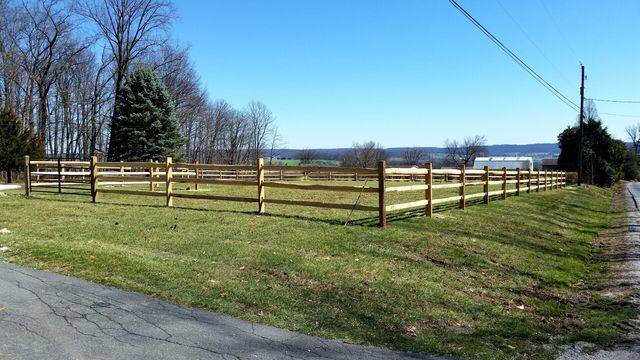 A wooden three-rail fence encloses a grassy field near a road on a sunny day with trees in the background.