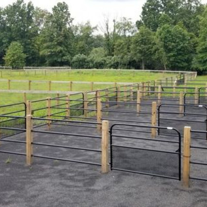 Black metal livestock fencing panels installed between wooden posts on a gravel surface next to a grassy pasture.