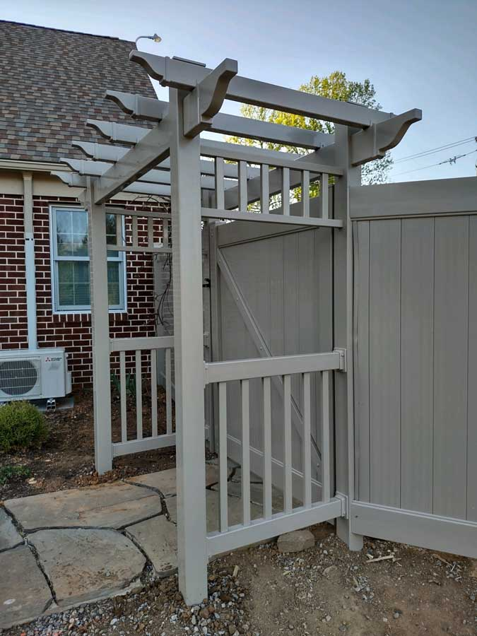 A light-gray wooden pergola entryway featuring vertical slats, standing on a stone path next to a matching privacy fence.