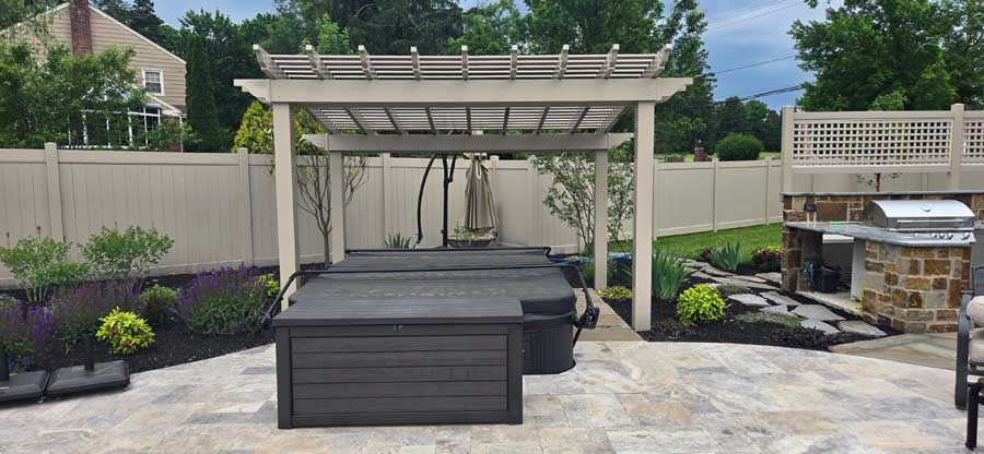 A backyard patio featuring a hot tub under a white pergola, with a stone outdoor kitchen nearby and a vinyl privacy fence.