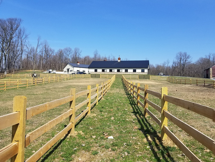A long, white horse barn with a black roof sits at the end of a path between two wooden rail fences in a grassy field.