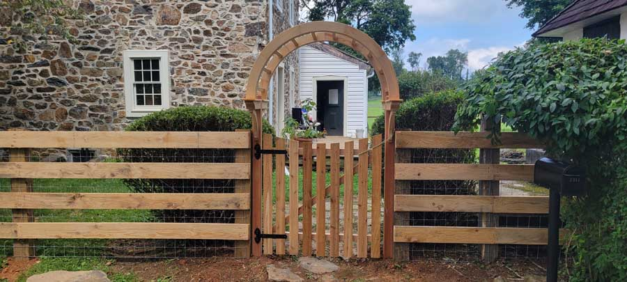 A wooden arched gate integrated into a horizontal slat fence in front of a stone house.