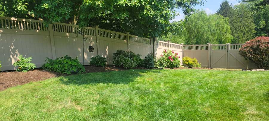 A grassy backyard with a beige vinyl fence, garden beds, and green trees under a clear blue sky.