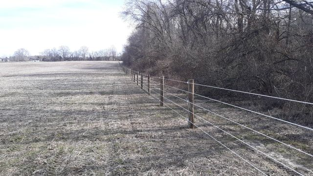 A wire fence with wooden posts runs alongside a field of dry, brown grass, bordering a line of leafless trees.