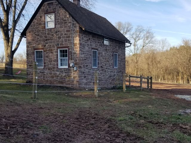 A small, two-story stone house with a dark roof stands in a field surrounded by a wire fence on a sunny day.