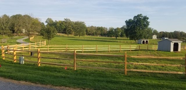A wooden rail fence borders a green grassy pasture with a small shed under a clear blue sky.