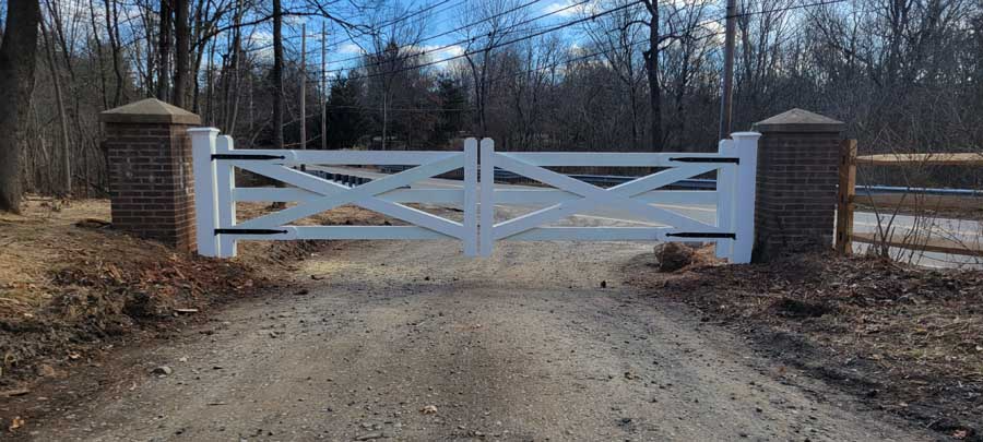 White wooden double gate with an X-pattern design mounted between two brick pillars on a dirt road.