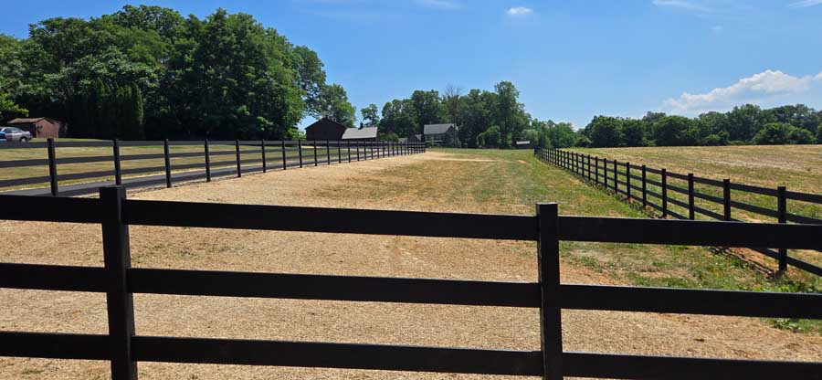 A wide view of a dry, grassy field enclosed by black wooden fences under a clear blue sky, with distant trees and buildings.