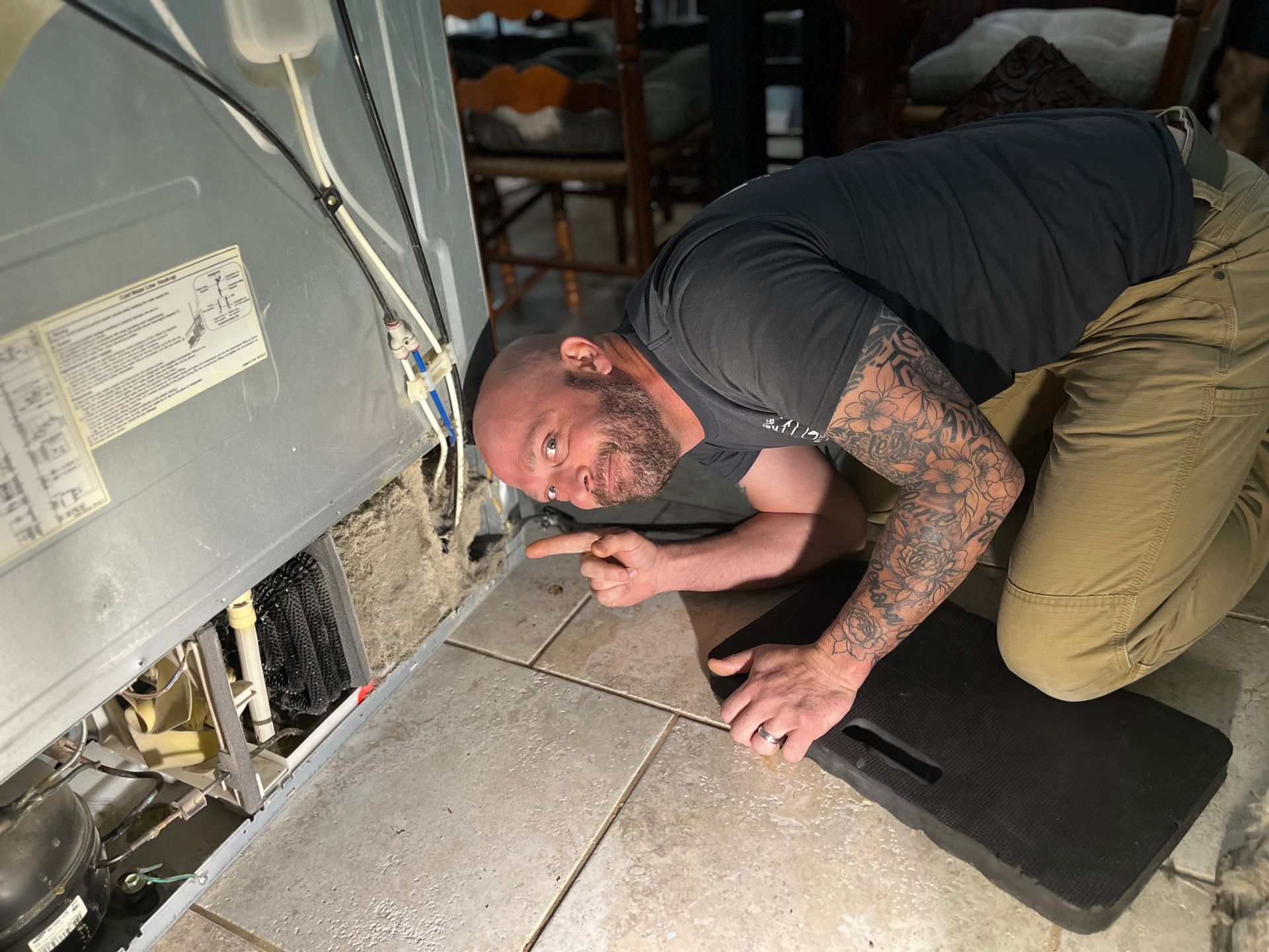 A man is kneeling on the floor looking under a refrigerator.