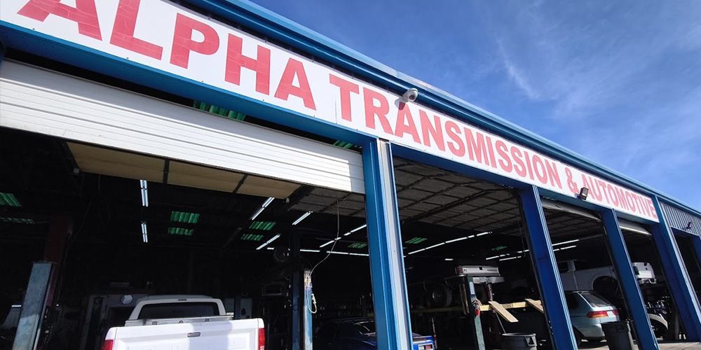 Auto repair shop with open service bays and a red “ALPHA TRANSMISSION” sign above the entrance
