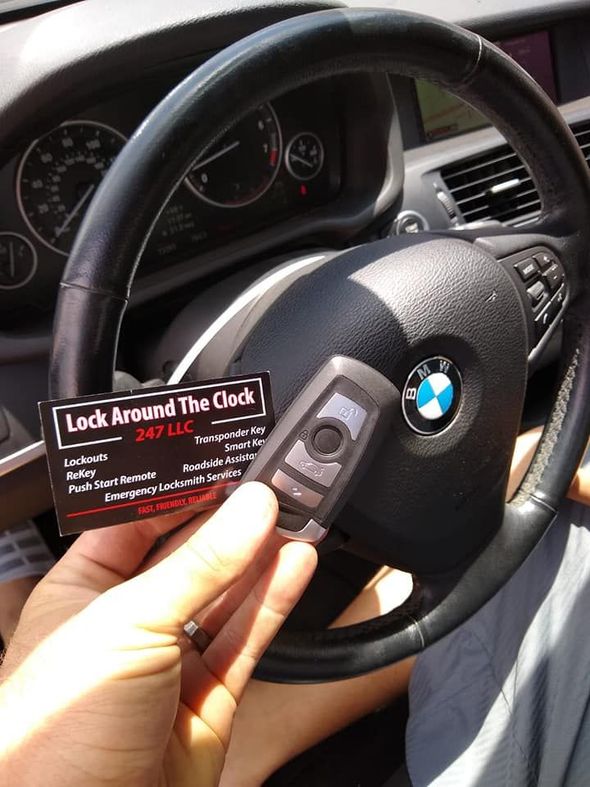 A hand holding a car key and a business card for Lock Around The Clock in front of a BMW steering wheel.