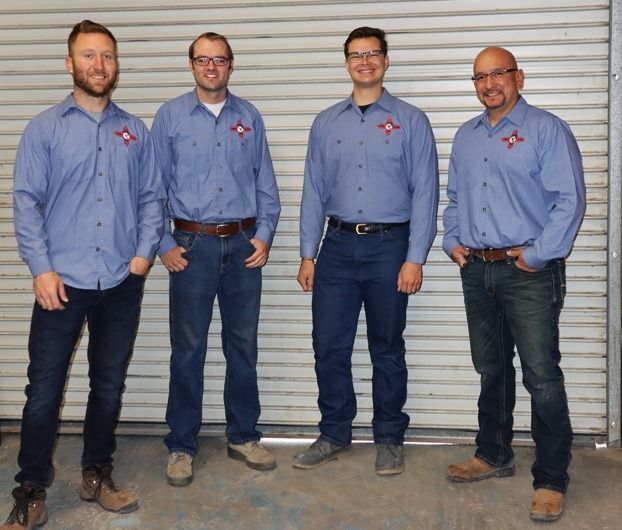 Four people in matching blue long-sleeved work shirts and jeans standing in a row against a closed metal roll-up door.