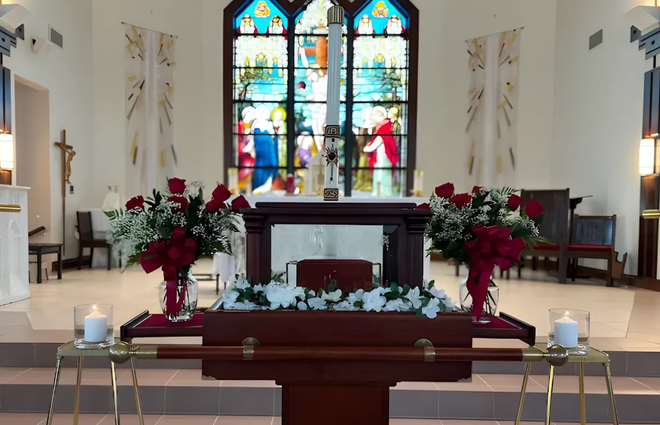 Church altar with red flowers, candles, and a stained-glass window behind it