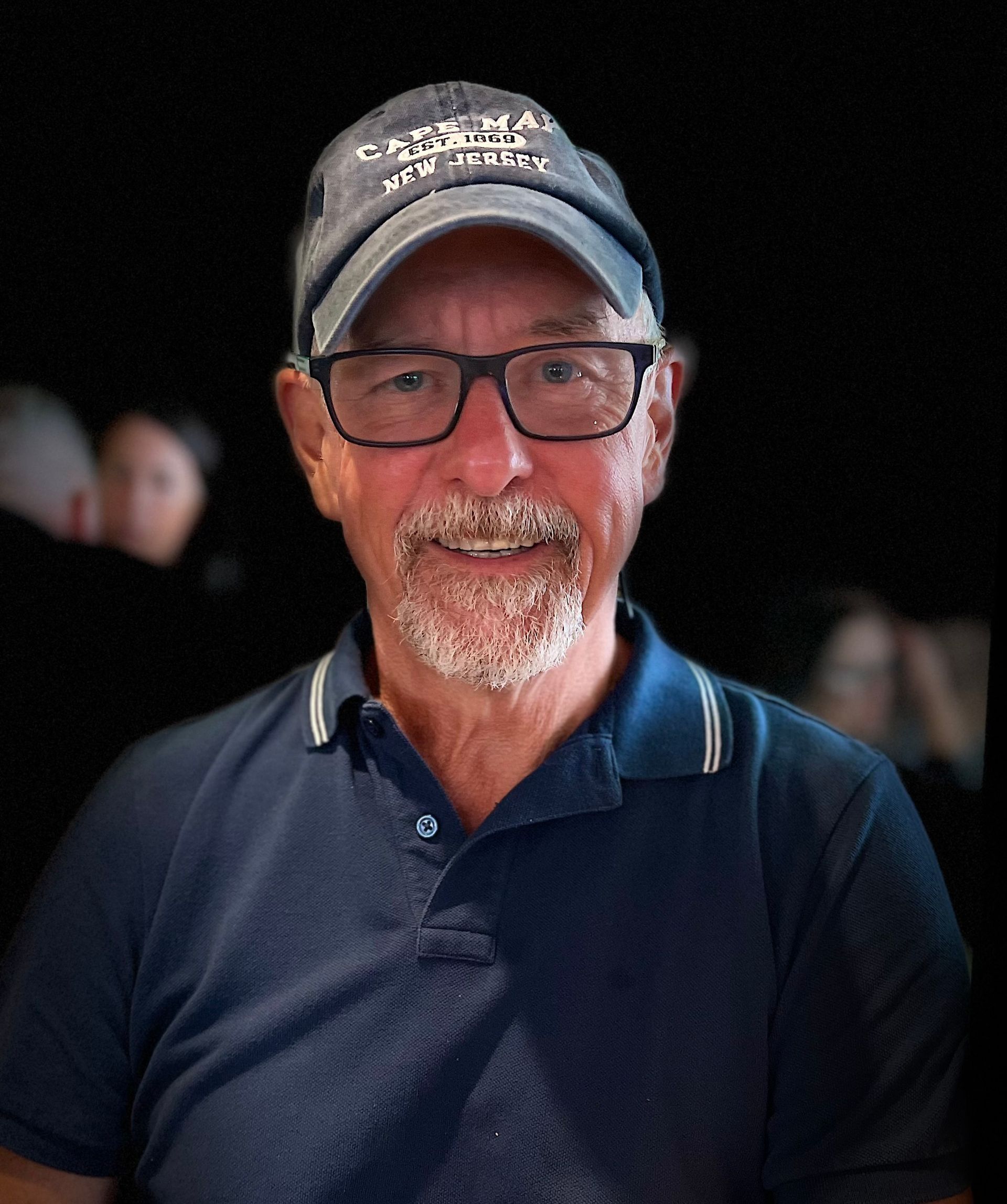 Man wearing glasses and a navy polo shirt, smiling in a dark background setting.