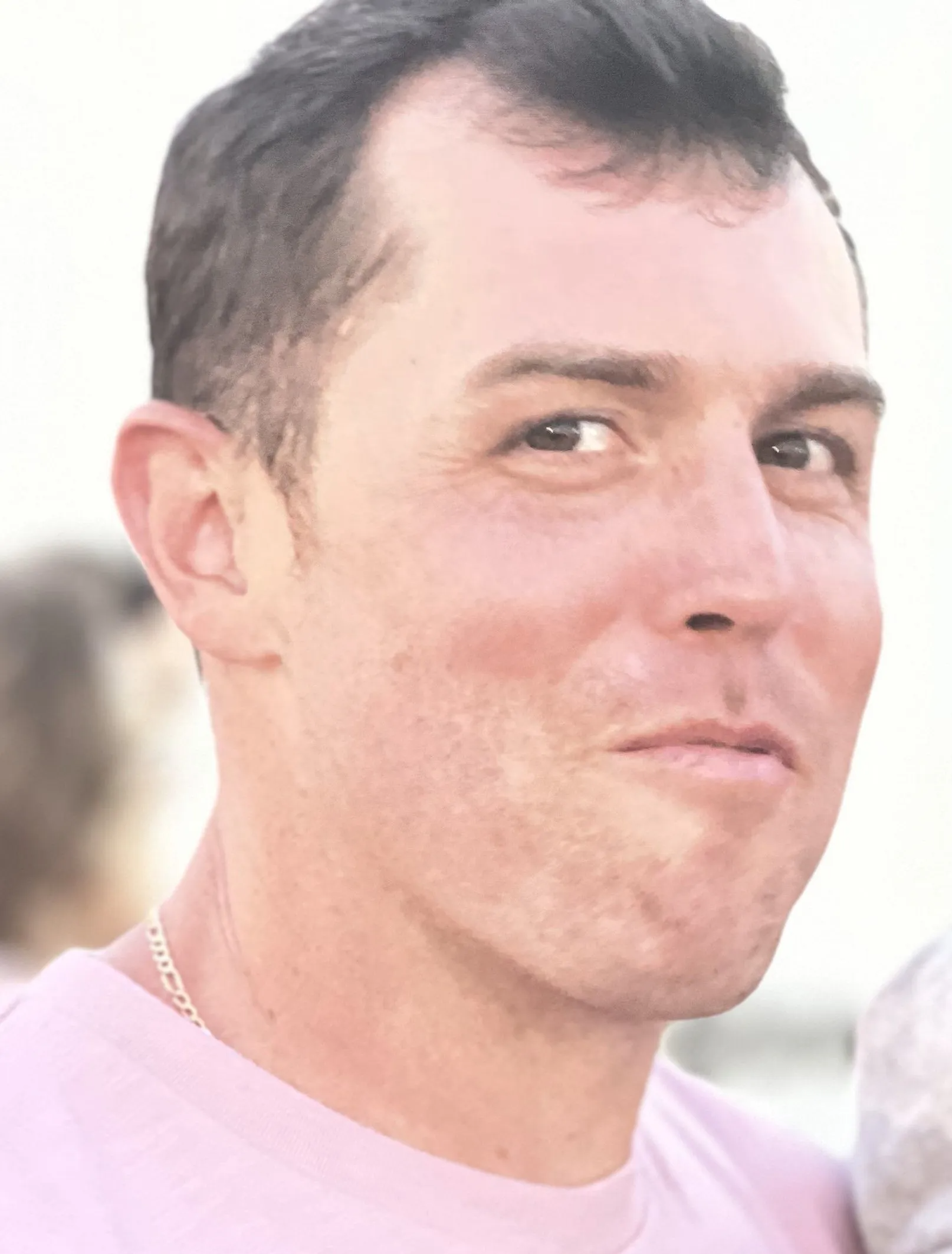 Close-up of a man in a light pink shirt, smirking slightly against a bright outdoor background