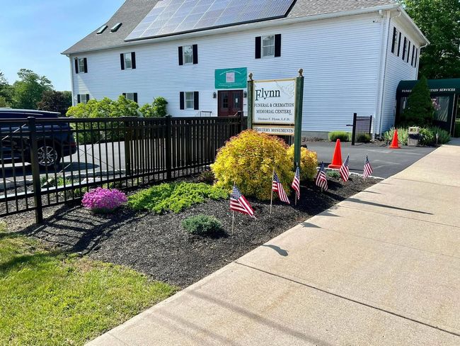 Sidewalk beside white building with black fence, flower beds, and orange cones on a sunny day