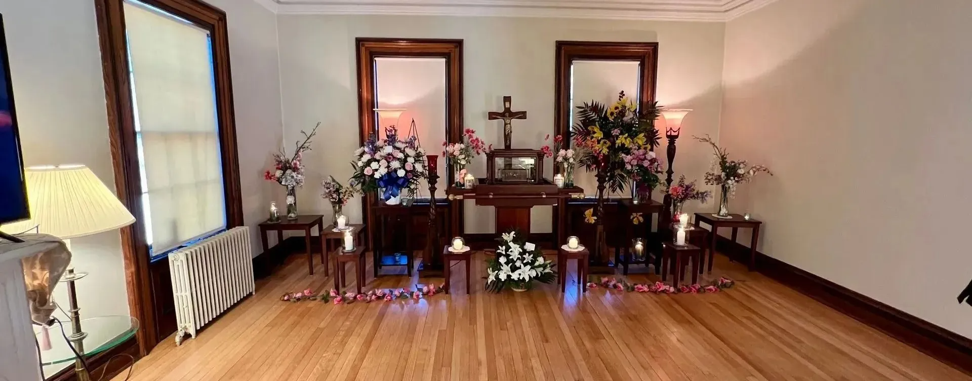 Memorial setup with framed photos, flowers, candles, and lights in a room with hardwood floors