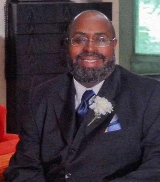 Smiling man in a dark suit with blue tie and white boutonniere, seated indoors