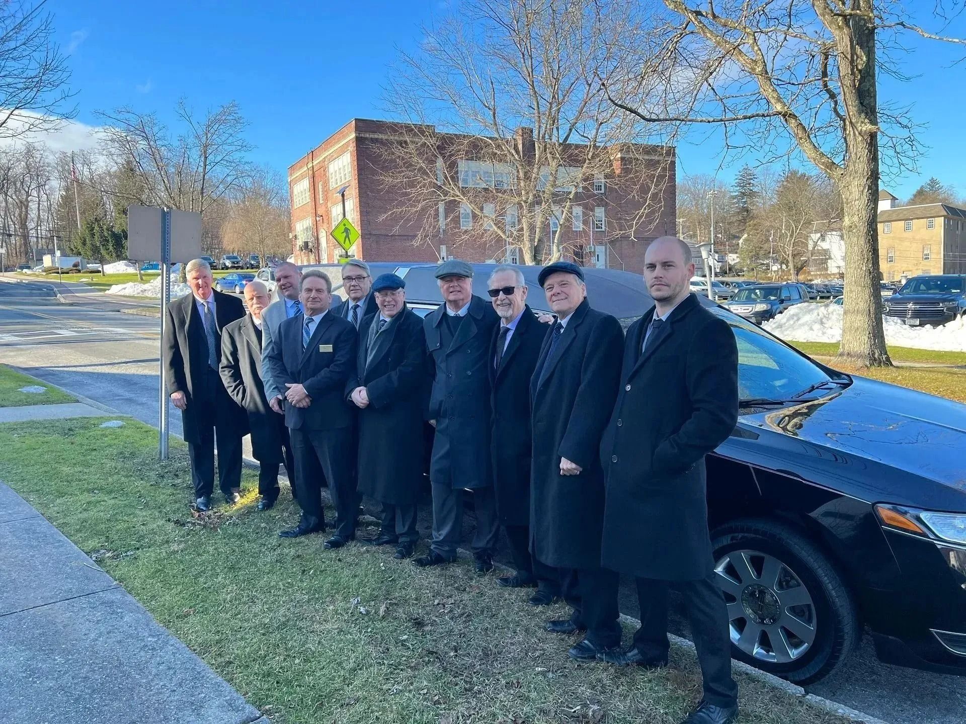 Group of people in dark coats standing on a sidewalk beside parked cars and a brick building on a sunny day