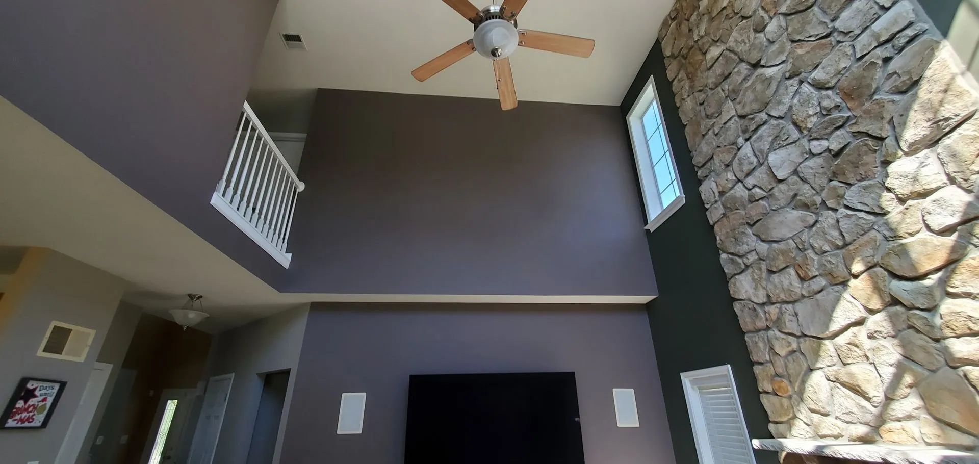 High-ceiling living room with stone wall, gray walls, ceiling fan, and bright windows