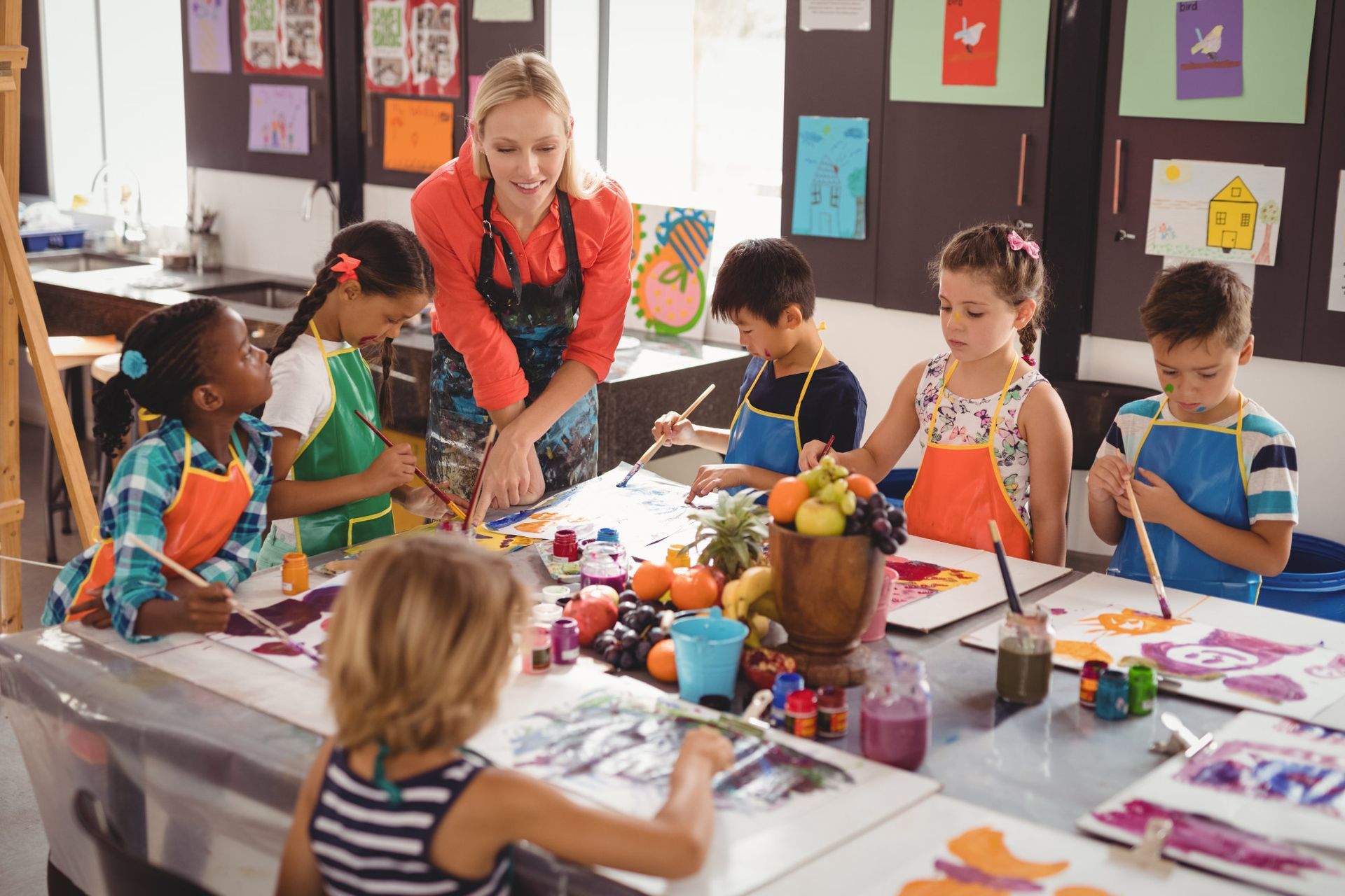 Une enseignante et des enfants peignent sur une table de classe avec du matériel d'art coloré et des dessins.