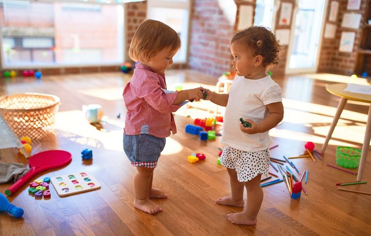 Deux tout-petits jouent avec des jouets colorés dans une salle de classe ensoleillée, tendant les bras l'un vers l'autre.