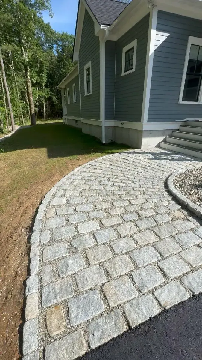 Stone path curves towards a house with blue siding. Lawn on the side. Sunny day.