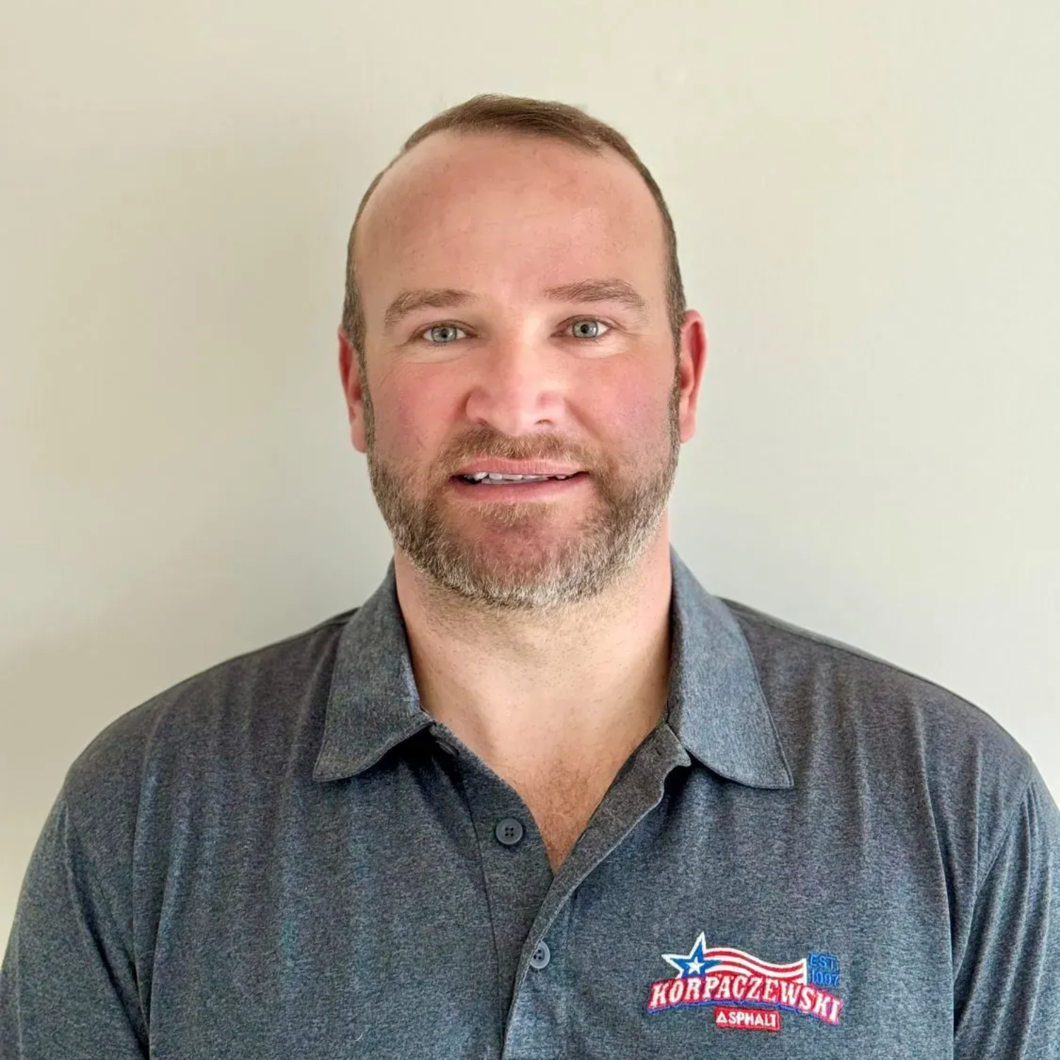 Man with a beard wearing a gray polo shirt with a logo, smiling, indoors.