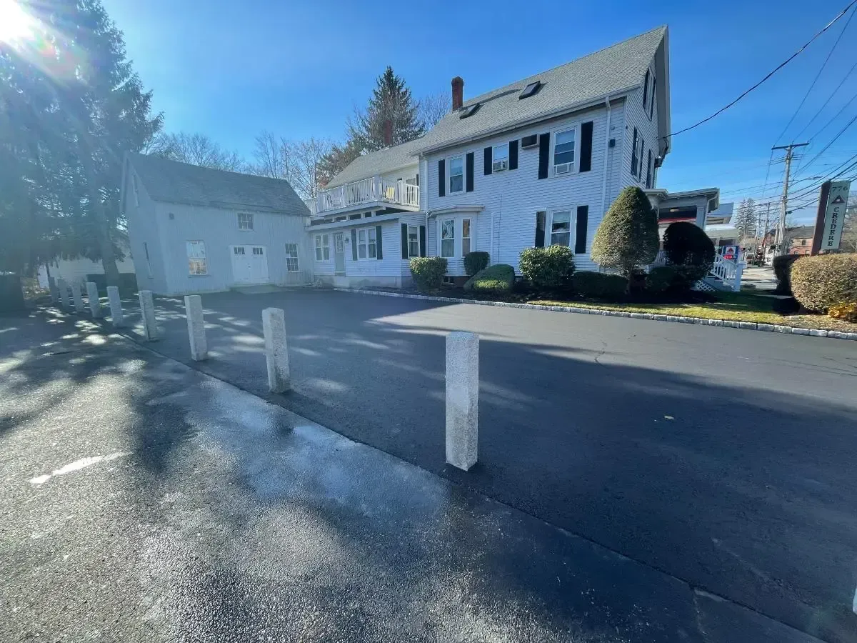 White building with black shutters and a driveway with concrete posts. Sunny day.