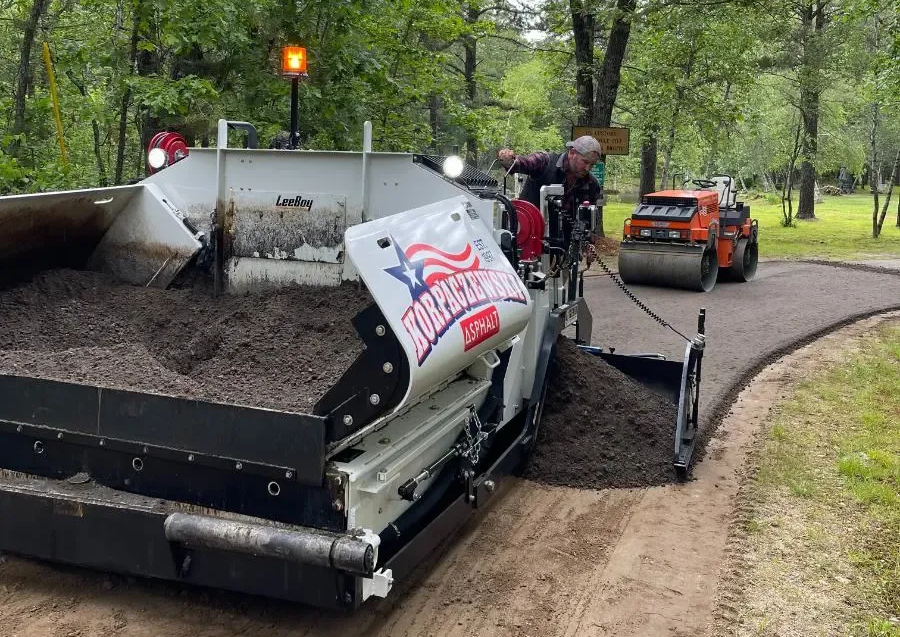 Asphalt paving machine laying asphalt on a road, worker operating it, roller in background.