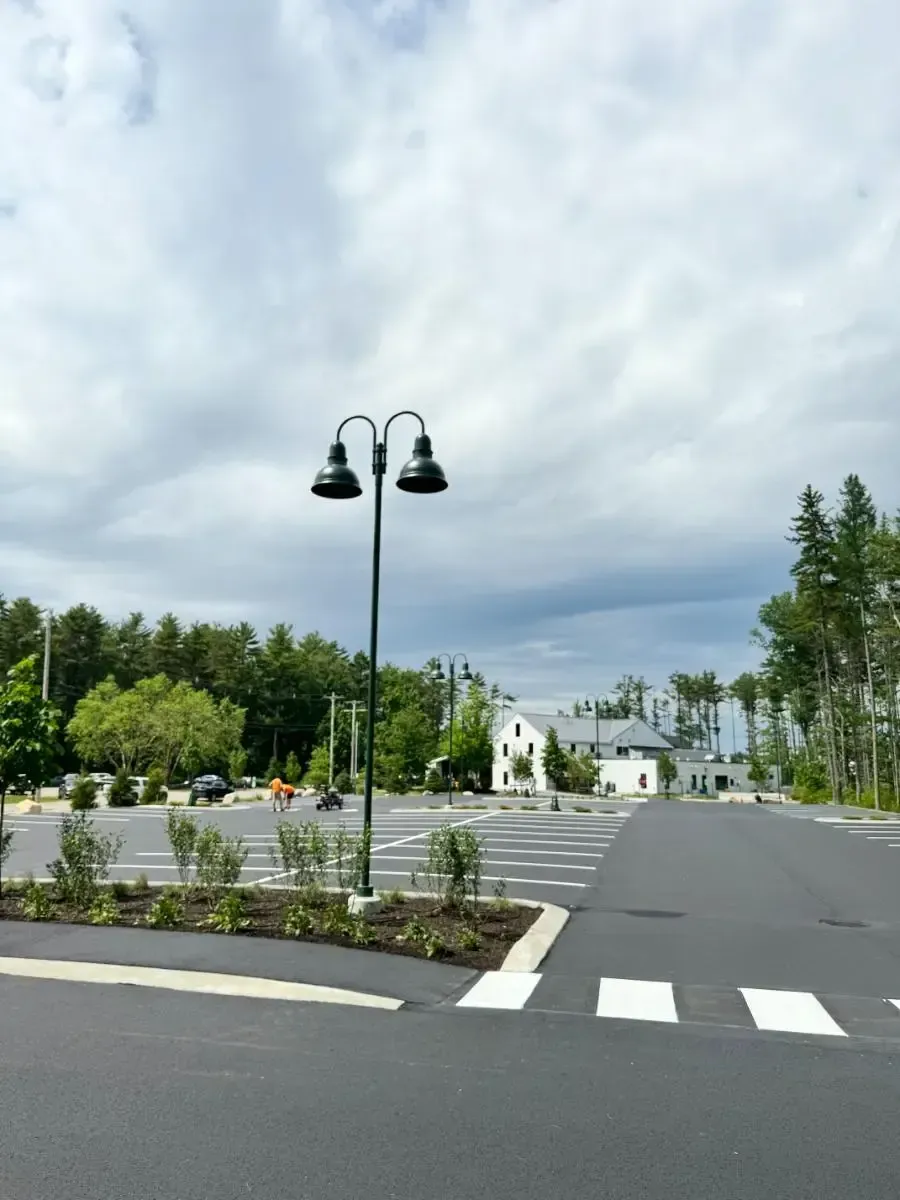 A paved parking lot with a street lamp, crosswalk, and white buildings against a cloudy sky.