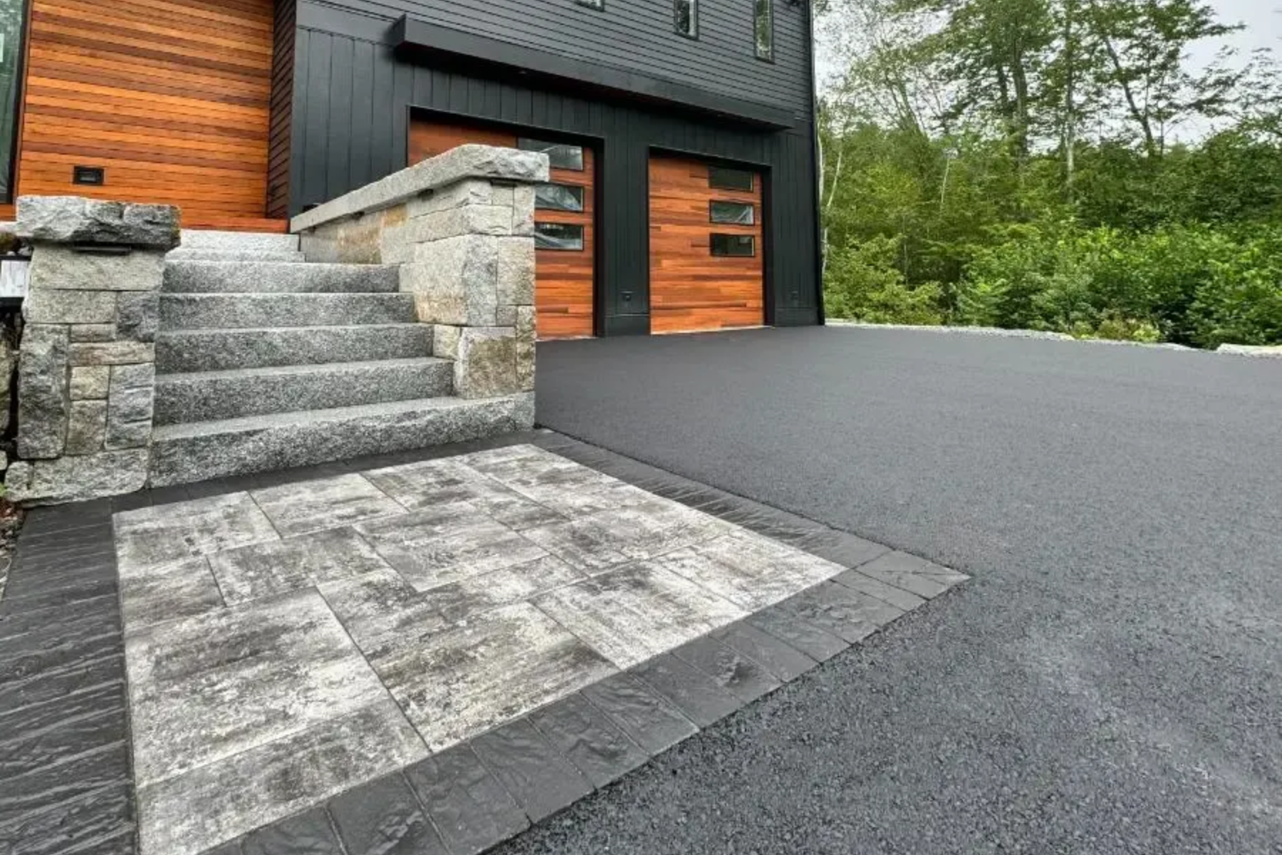 Stone steps and walkway leading to a modern home with black siding, wooden garage doors, and asphalt driveway.