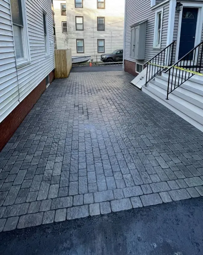 Brick paver driveway between two buildings. One side is a house, the other an entrance with stairs.