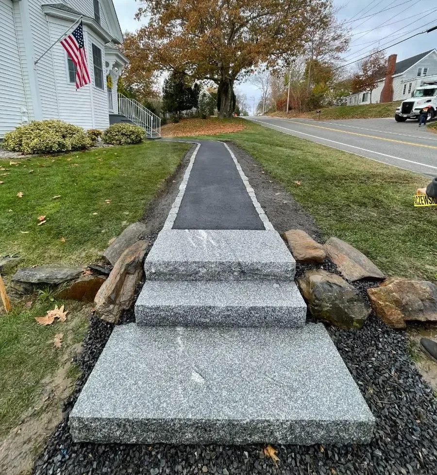 Granite steps and walkway lead to a road, passing a house with an American flag.