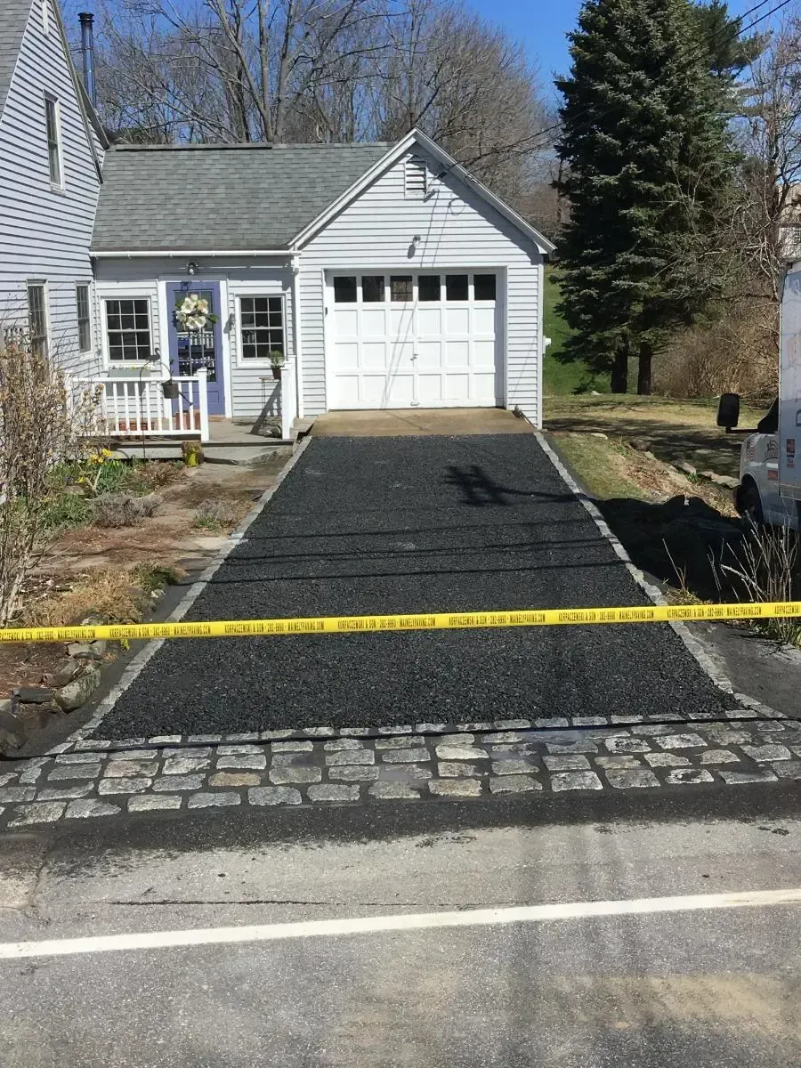 Newly paved asphalt driveway leading to a white garage, bordered by cobblestones.