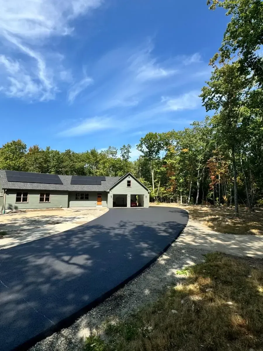 House with teal siding, dark driveway, and green lawn under a blue sky, framed by trees.