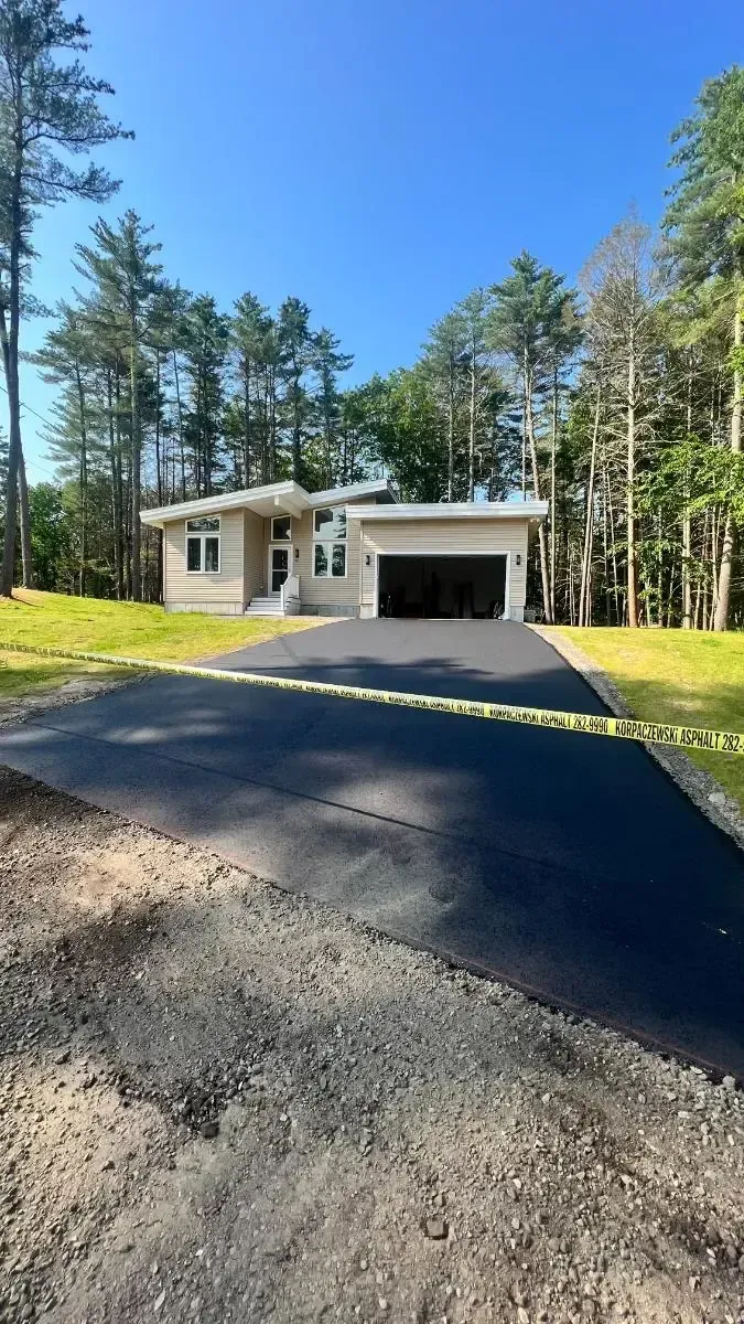 Modern beige house with black garage, new asphalt driveway, and surrounding trees under a blue sky.