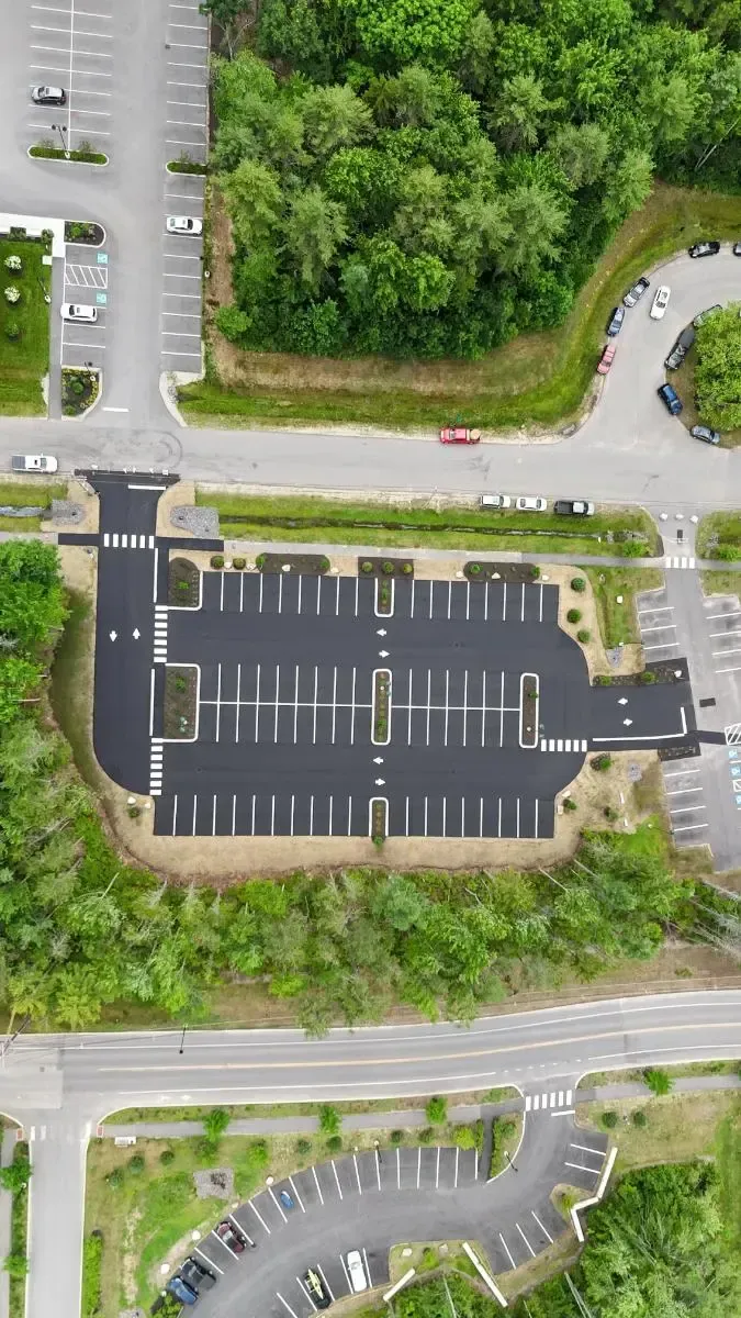Aerial view of a large asphalt parking lot surrounded by trees, with cars parked in designated spaces.