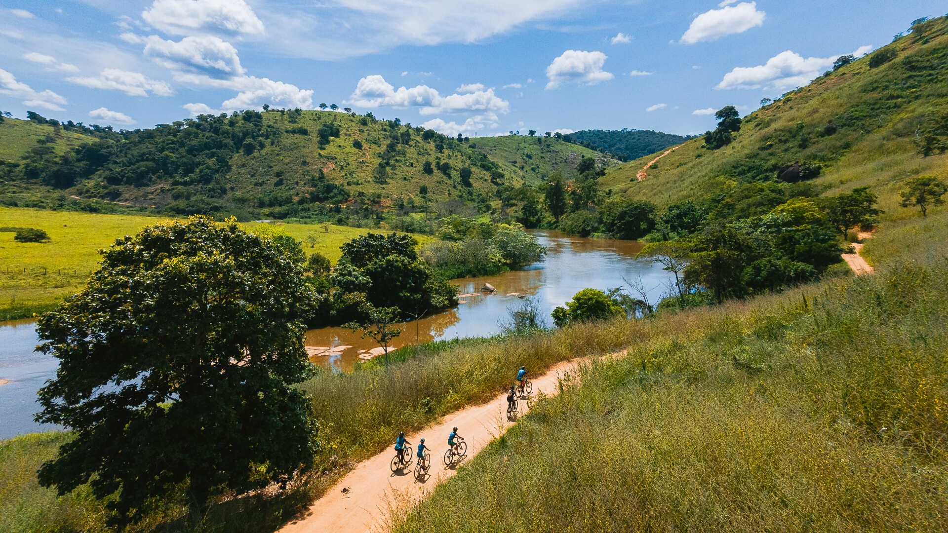 Um grupo de pessoas anda de bicicleta por uma estrada de terra próxima a um rio.