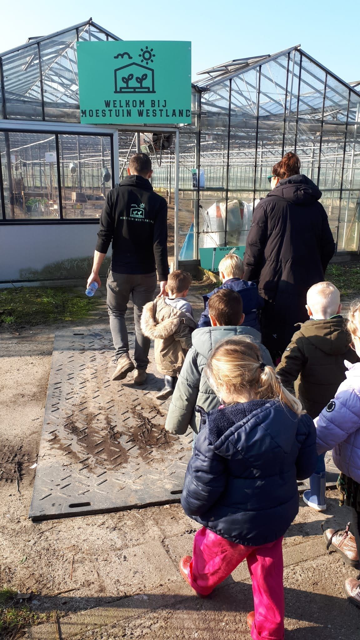 Kinderen volgen volwassenen in de buurt van een kas met een bordje: zonnige dag.