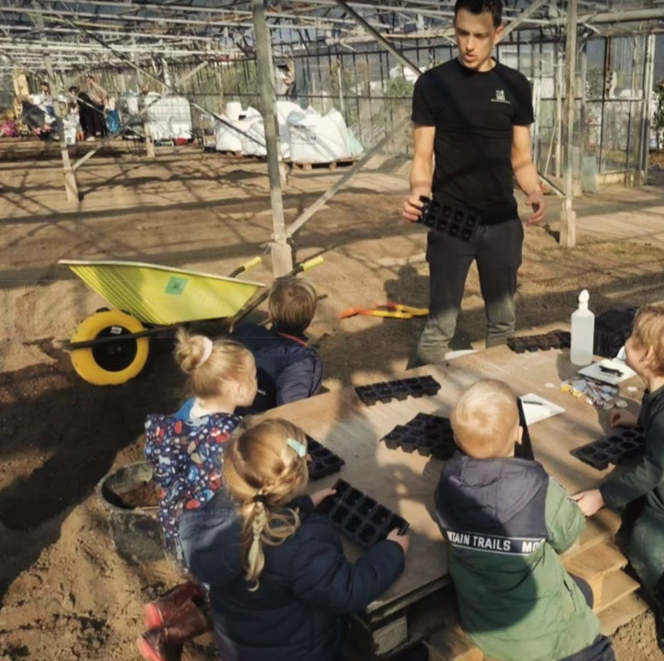Een man leert kinderen in een kas hoe ze moeten planten. Ze houden zaaibakjes vast.