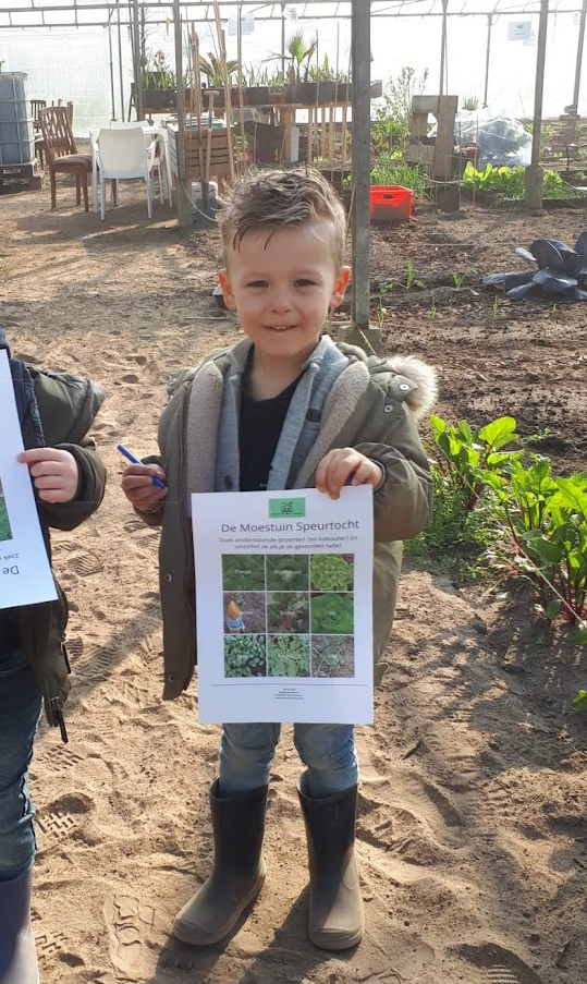 Een jongen in een kas houdt een vel papier met afbeeldingen van planten vast, draagt ​​laarzen en een jas en lacht.