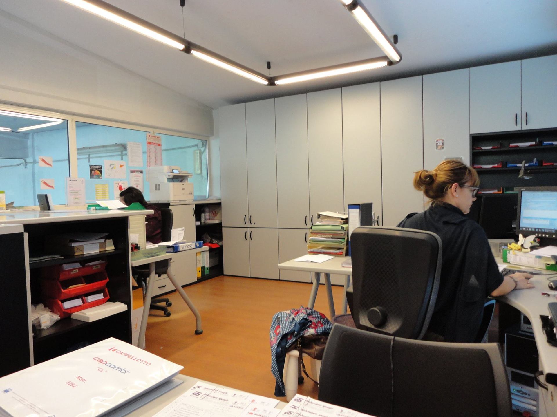 Interno di un ufficio con una persona che lavora a una scrivania, schedari e una finestra.
