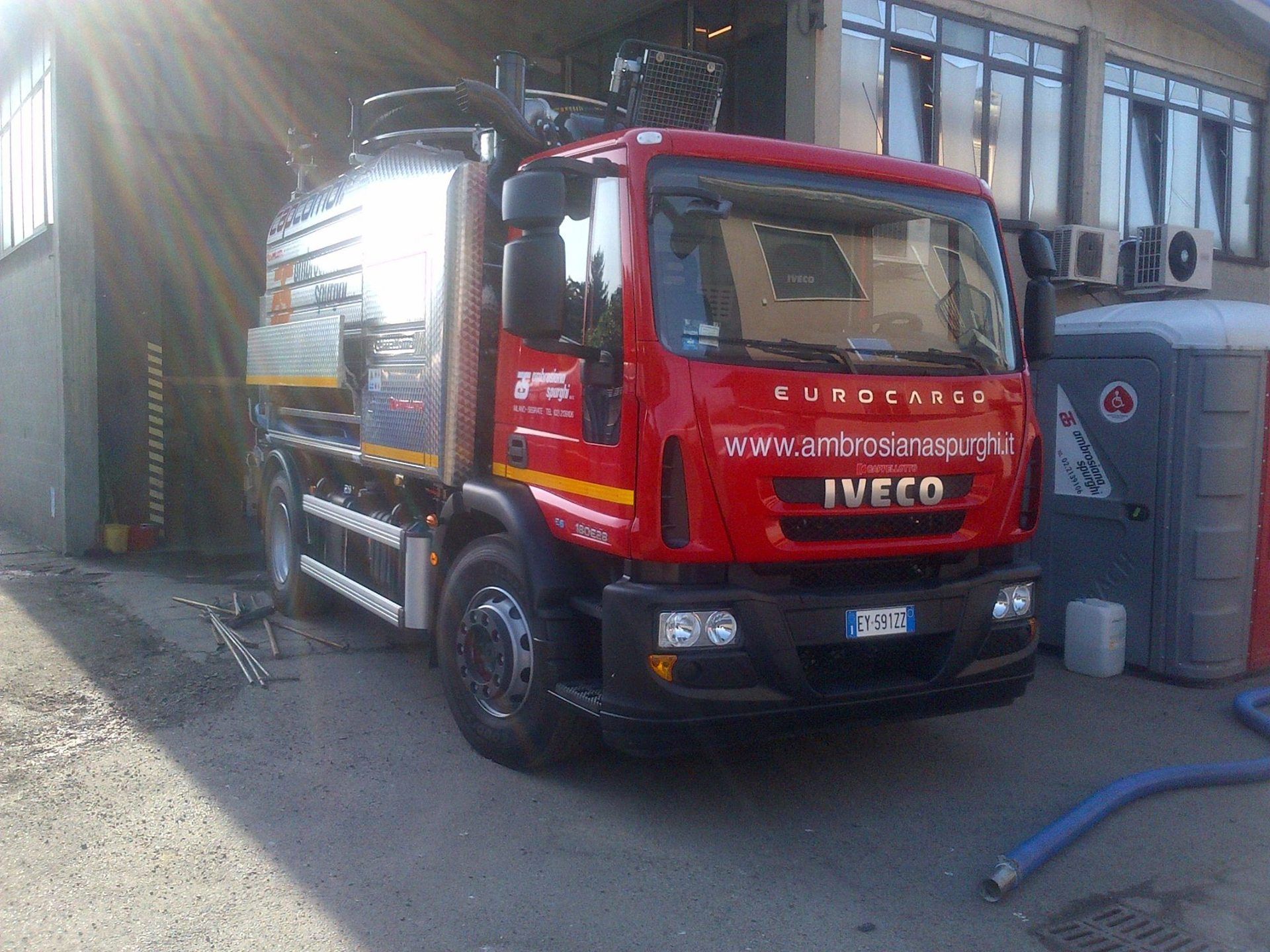 Un camion Iveco rosso con cisterna, parcheggiato vicino a un edificio con bagni chimici.