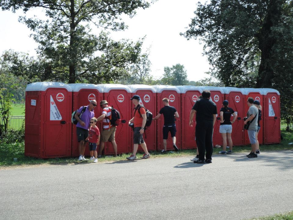 Bagni chimici rossi allineati. La gente aspetta di usarli su un sentiero vicino agli alberi.
