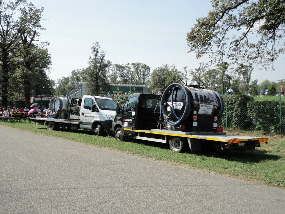 Due camion con rimorchi che trasportano grandi oggetti cilindrici neri parcheggiati su una strada, con alberi e un campo verde sullo sfondo.