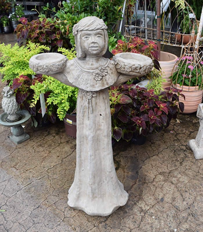 Statue of a child with outstretched arms holding two bowls, likely for plants. Outdoors, surrounded by greenery.