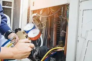 HVAC technician using gauges to service air conditioning unit with exposed wires and copper tubing.